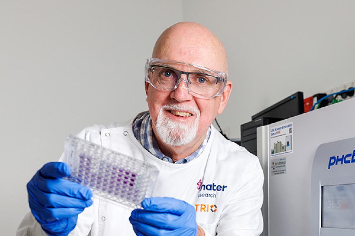 a man in a white coat, glue gloves and glasses holds up a small tray of purple dots