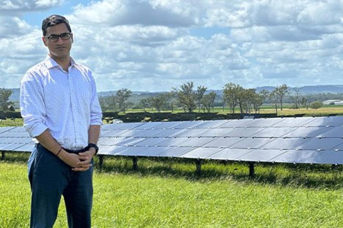  A man stands in a grassy field with rows of solar panels behind him. 