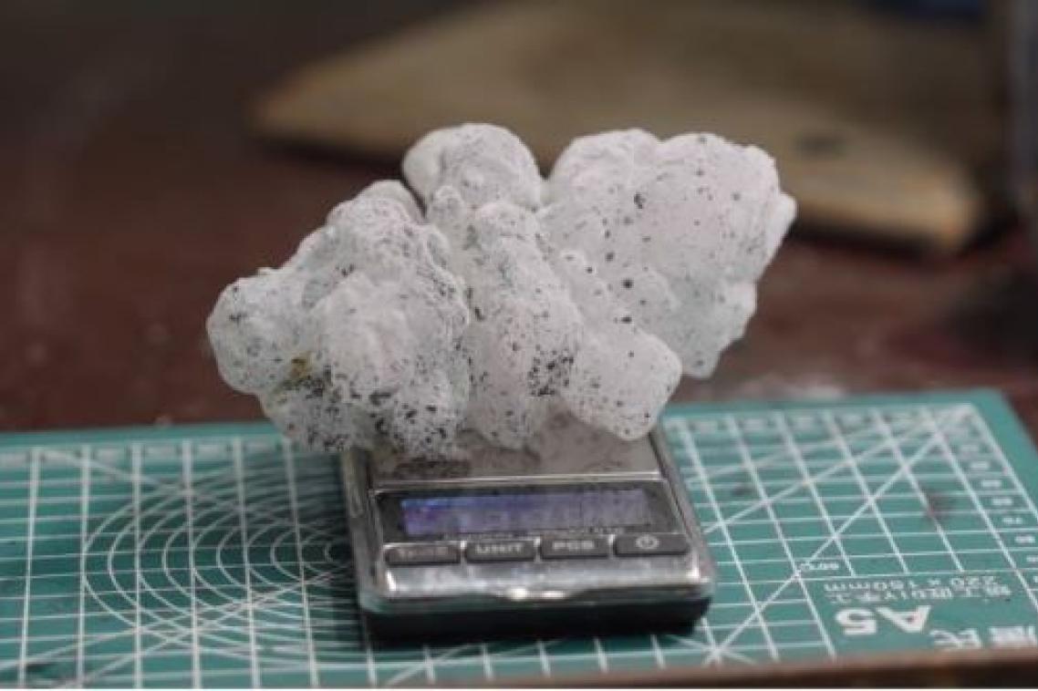 A spiky white hailstone, flecked with small dots of black paint, rests on a scale on a desk.