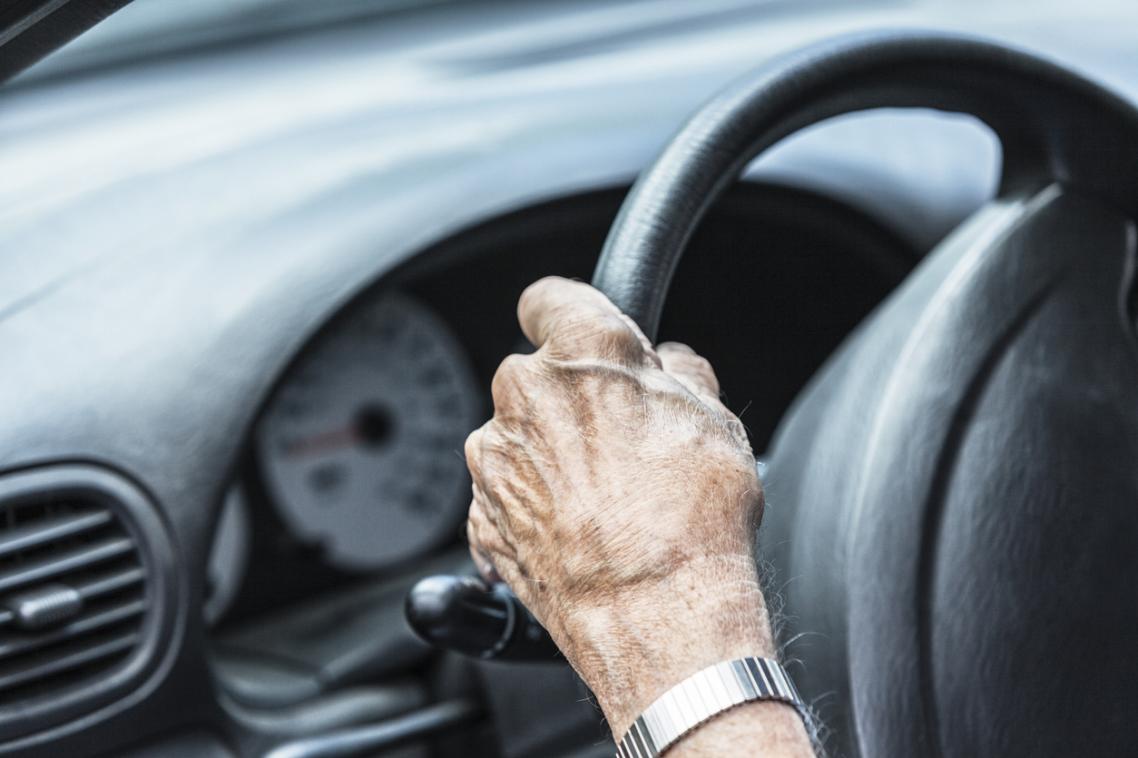 A older person with their hand on a car steering wheel.