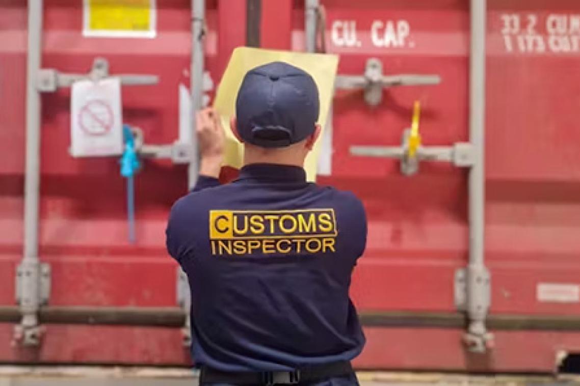 a man in a blue shirt with Customs Inspector on it looks at a red shipping container