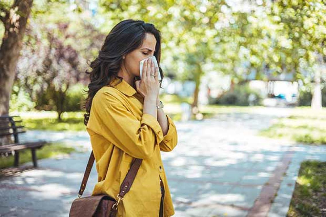 Woman in yellow jacket blowing her nose in a tissue standing in a city park with trees in the background