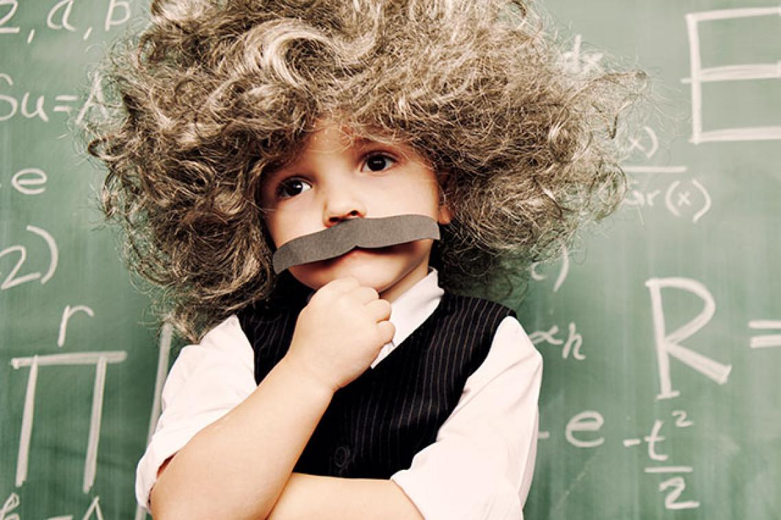 A young child dressed as Einstein stands in front of a chalk board covered in equations