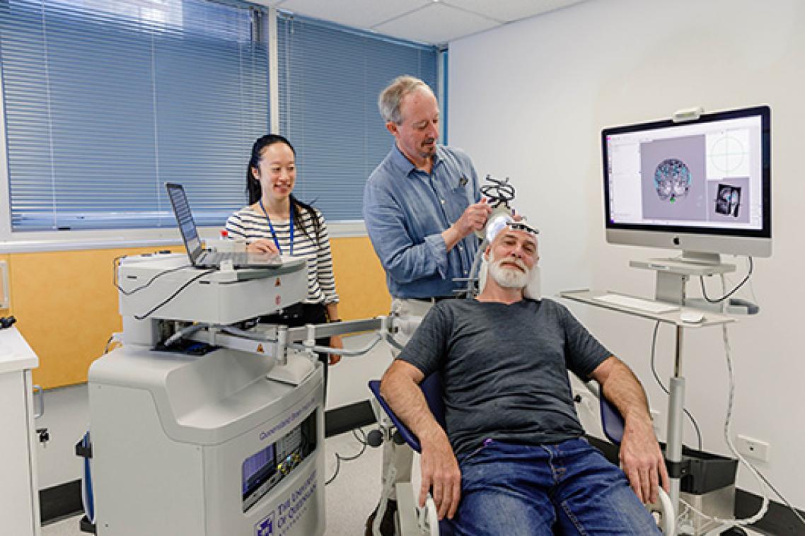 A man sitting on a chair receiving ultrasound treatment on his head in a lab. Two ultrasound technicians are behind him.